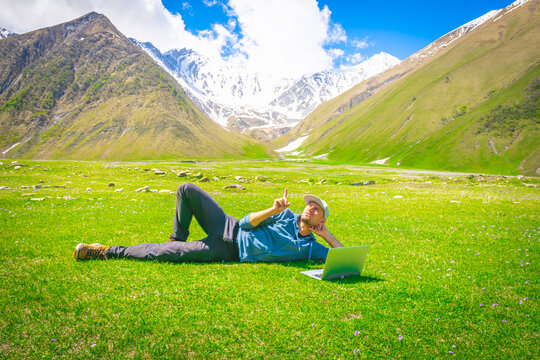 Male person in csualclothes  lays on the grass and points his finger up with mountains in the background. Inspiration and new ideas born in nature concept. Backgorud image of creativity