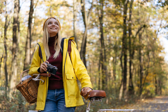 Smiling photographer holding retro camera, planning taking pictures in park. Beautiful woman wearing yellow raincoat, hipster hat standing near bicycle in autumn forest. Inspiration, travel concept
