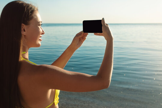Girl In Swimsuit Holding Phone Taking Picture Of Sea Outdoors