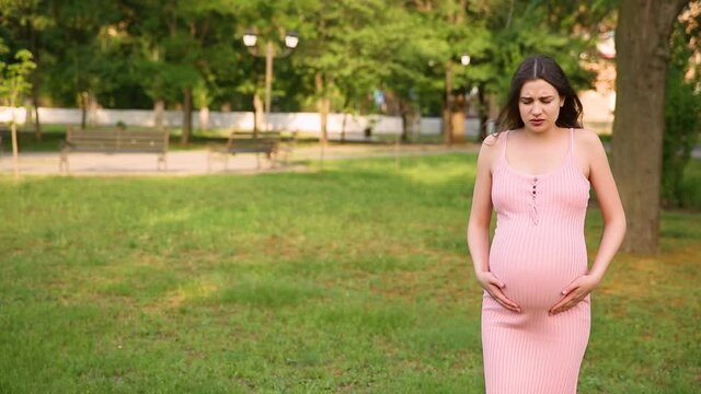 A pregnant girl is standing in the park, and her stomach begins to hurt. a pregnant woman strokes her stomach and walks from side to side.