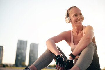 Fototapeta na wymiar Young caucasian woman athlete sitting and relaxing after workout and physical exercise. Sporty girl listening music in headphones. Backlit sunset, city view. healthy lifestyle, motivation concept