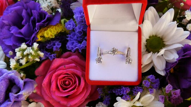 Gift present red box with a set of engagement ring and earrings on the background of a multi-colored beautiful bouquet of flowers. Marriage proposal. White gold or silver with diamond stone