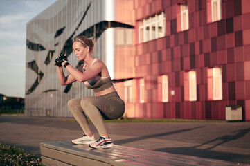 Woman athlete is jumping on bench in park. Sporty girl is doing physical exercise outdoor