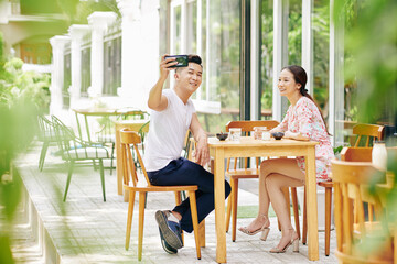 Handsome young Vietnamese man taking selfie when eating breakfast with girlfrind in outdoor cafe