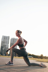 Fototapeta premium Fitness woman taking a break after a workout, strong sporty girl in headphones stretching outdoors, back lit sunset, city view