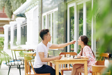 Caring young Vietnamese man giving girlfriend a spoon of fresh yogurt he ordered in cafe
