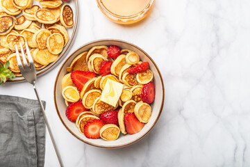 Trendy home breakfast with tiny cereal pancakes with piece of butter and fresh strawberries in the bowl, grey napkin and honey nearby