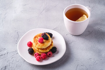 Cottage cheese pancakes, curd fritters dessert with raspberry and blackberry berries in plate near to hot tea cup with lemon slice on stone concrete background, angle view