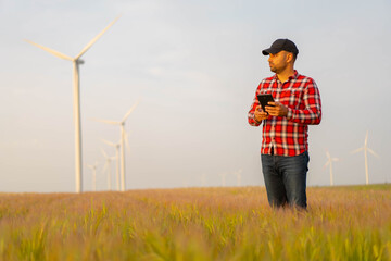 Male farmer standing in wheat field with tablet  inspecting crops. Wind mill in background