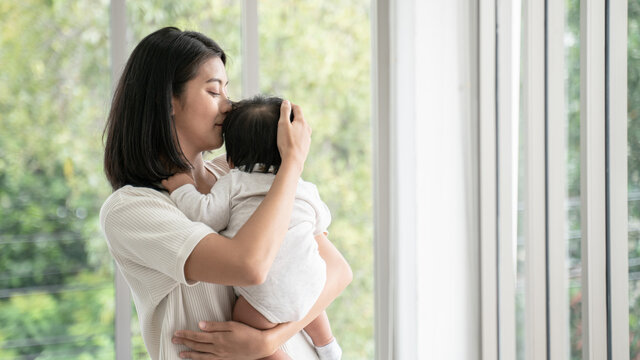 Close up of asian beautiful mom hugging sleeping newborn baby in her arm and kissing the kid.Asian mother look out the window and holding her baby head in home.she is happy to stay at home with baby.