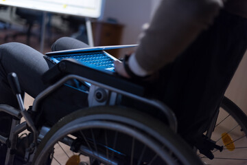 Disabled businessman in the wheelchair works in the office at the computer while performing in co-working space. Disability and handicap concept. Horizontal shot. Selective focus.