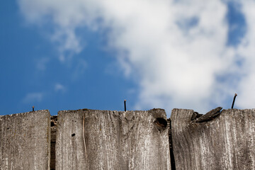 Poszarzałe od słońca stare deski, z wystającymi gwoździami. Fragment ruiny drewnianego wiejskiego domu, w tle nieostre błękitne niebo z chmurami. © AP-STUDIO