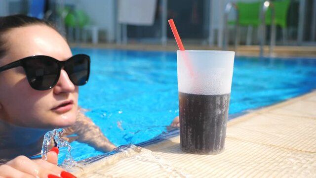 Close up glass of cool cocktail standing near pool. Girl swim on edge of basin to refresh herself with cool drink. Attractive woman relaxing on luxury resort with cold alcohol beverage. Slow motion