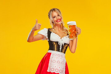 Young sexy Oktoberfest girl waitress, wearing a traditional Bavarian or german dirndl, serving big beer mug with drink isolated on yellow background. Wow emotion, woman with thumbs up.