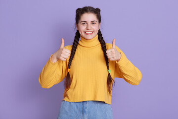 Portrait of beautiful cute girl standing with pigtail hairstyle in yellow shirt, woman posing isolated over lilac background, excited girl looks at camera with charming smile.