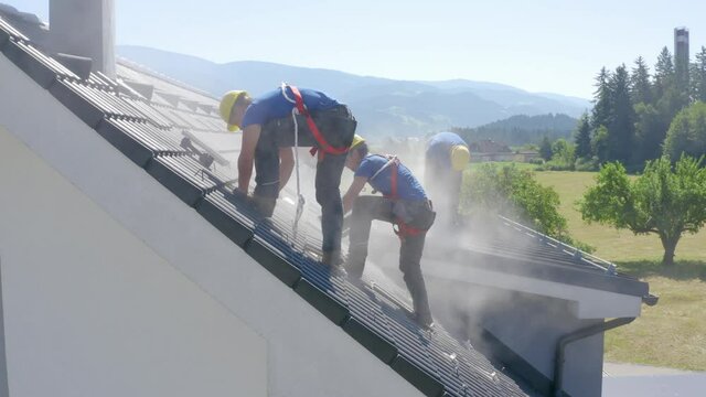 Three young male solar power roof installers with yellow hard hats, blue shirts, and safety harness work and stand on dusty residential rooftop with mountain range in background, aerial