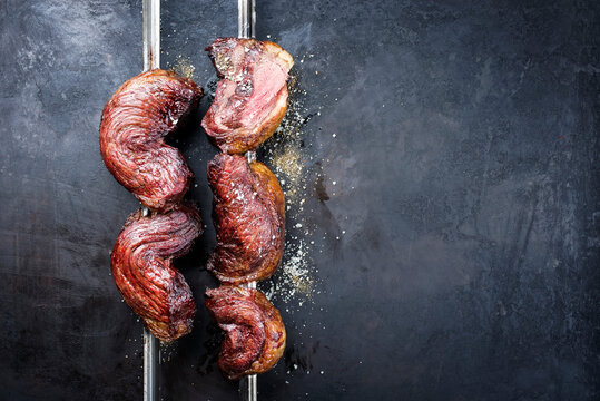 Gegrillte brasilianische dry aged Wagyu picanha Grillspie&szlig;e H&uuml;ftdeckel vom Rind angeboten als Draufsicht auf einem rustikalen schwarzen Board mit Textfreiraum rechts