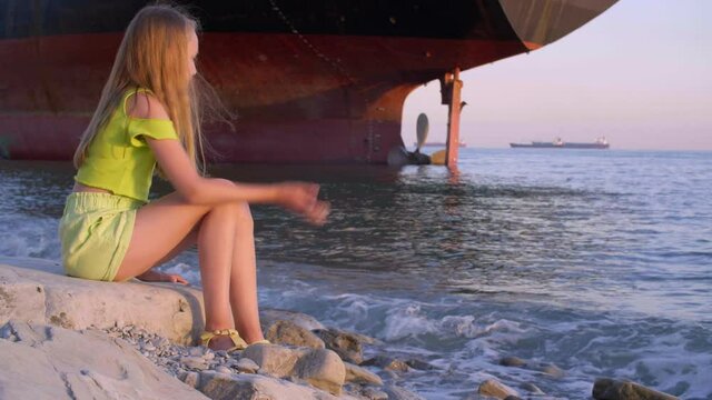 Traveling girl teenager throwing stones to sea water on cargo ship landscape on horizon. Tourist girl throwing rocks to water on sea shore on freight ship background