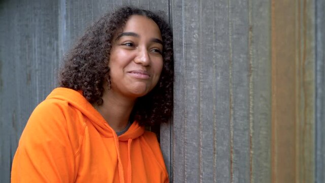 Beautiful mixed race African American girl teenager young woman looking sad then laughing and happy wearing an orange hoodie in an urban city environment