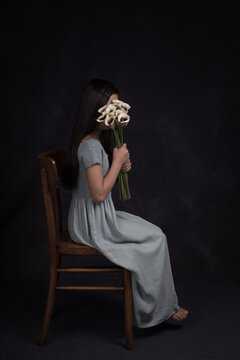 Studio portrait of an anonymous young girl in long blue dress sitting on a chair with bouquet of  white with purple arums in front of her face
