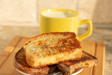 Egg-fried croutons of white and black bread. In the background is a yellow cup with milk. Illuminated by bright daylight.