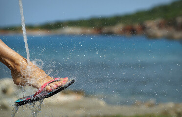 Foot in beach slides under water spray, water drops splashing around. Blue sea and coastline. Free...