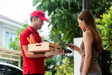 Naklejka na ściany i meble Delivery staff on red uniform holding box of fresh food delivering to customer's home and paid by online transaction use smartphone