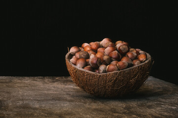 Coconut shell bowl full of hazelnuts in the shell on a wooden table on black background