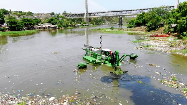 Green dredger cleaning rubbish in a polluted brown river, bridge and poor population living on the river bank