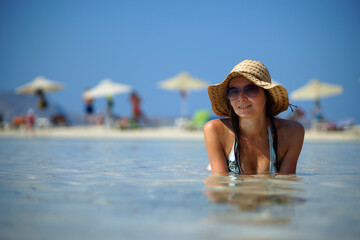 Young smiling woman in bikini, straw hat and sunglasses lying in shallow water. White sand and sun umbrella background