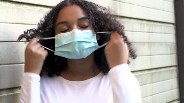 Beautiful mixed race African American girl biracial teenager young woman outside wearing and taking off a face mask during COVID-19 Coronavirus pandemic