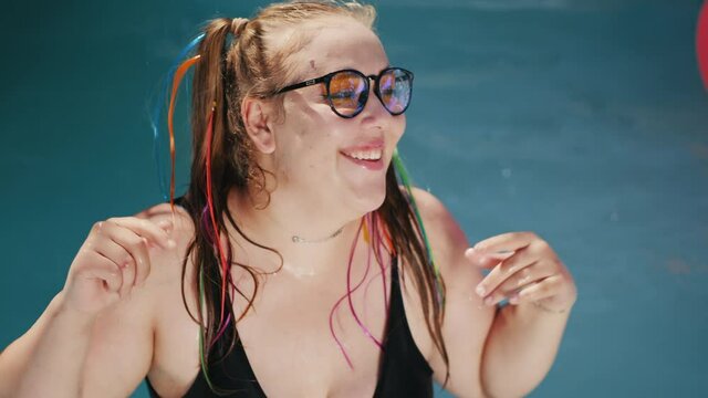 Body positivity. A plus-size young girl in a swimsuit and sunglasses dances in the middle of the outdoor swimming pool, lady splashes water and smiles.
