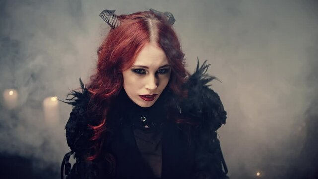 Ritual of Occultism or esoterics concept. Red-haired girl witch with black horns sitting on floor in candlelight and praying with folded hands. Scary demon doing witchcraft. Halloween mystery rite.