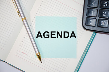 The word Agenda is written on a paper blue block near a calculator and notepad. pen next