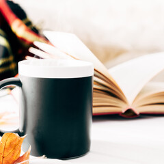 Autumn cozy composition. Plaid, steaming cup of hot chocolate, book, autumn leaves. Autumn or winter concept of coziness and interior. Front view, copy space, square