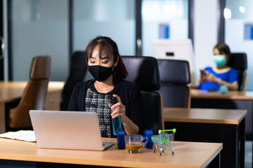 Young Asian woman officer disinfectant spray on Wipes cleaning laptop computer on desk office against corona virus COVID-19
