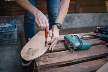 Man's hands making skateboard and using screwdriver. Cropped man assembling skateboard on table with electric saw in workshop.