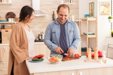 Man slicing tomatoes for salad on cutting board in kitchen while wife is smiling. Happy in love...