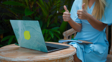 A girl in a blue dress remotely online working behind a laptop showing her thumbs in the backyard with green sprouts on the background