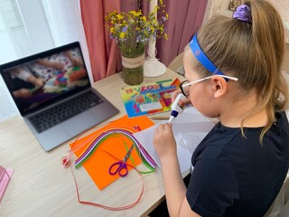 The girl in glasses makes handmade crafts. Glue and colored paper in the hands of a child. Items for creativity are on the table.