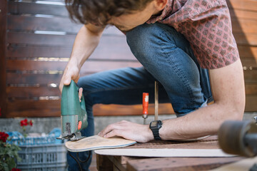 Cropped man using electric saw while making skateboard. Close-up view of male hand holding power saw and cutting wooden board in workshop.