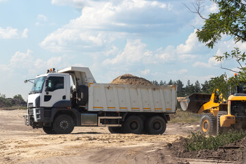 Dump truck with a bunch of land in the back and excavator at the construction site outside the city. Road works on an intercity highway on a cloudy day
