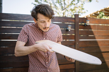 Man blowing wood dust from polished skateboard deck outdoor. Brunette man cleaning wooden board...