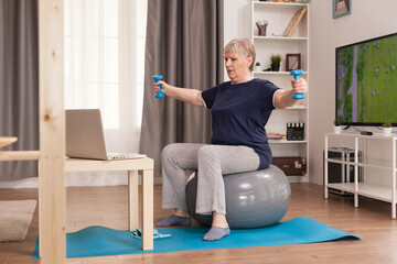 Retired woman training shoulders with weights. Old person pensioner online internet exercise training at home sport activity with dumbbell, resistance band, swiss ball at elderly retirement age