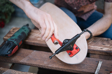 Close-up view of male hands assembling trucks on skateboard deck. Cropped man putting trucks on deck for making skateboard on wooden table.