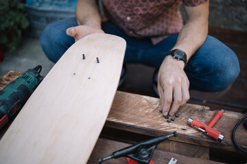 Wooden skateboard deck with bolts in male's hands. Man's hands screwing bolts into wooden board for making skateboard.