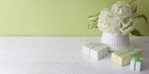 Little green gifts and a bouquet of white hydrangea in a vase. Green wall and white wooden table.