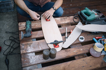 High angle view of man fitting truck on wooden skateboard. Male hands making skateboard while adding wheels to wooden deck.