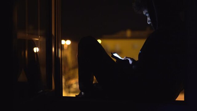 Young man confined with mobile phone sitting at window at home at night. Stock video of person in quarantine to prevent coronavirus infection.