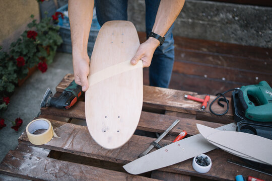 Cropped man attaching paper tape to skateboard. Man's hands sticking paper tape to wooden board for painting skateboard pattern.